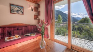 Ein Zimmer mit Holztisch, rotem Sofa, Blumenvase, gerahmtem Bild an der Wand und Balkon mit Bergblick.