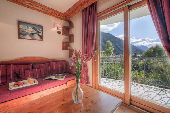 A room with a wooden table, a red sofa, a vase with flowers, a framed picture on the wall, and a balcony with a mountain view. - Résidence Le Cristal - Nant Blanc 4, Argentière (Chamonix), France (Argentière (Chamonix))