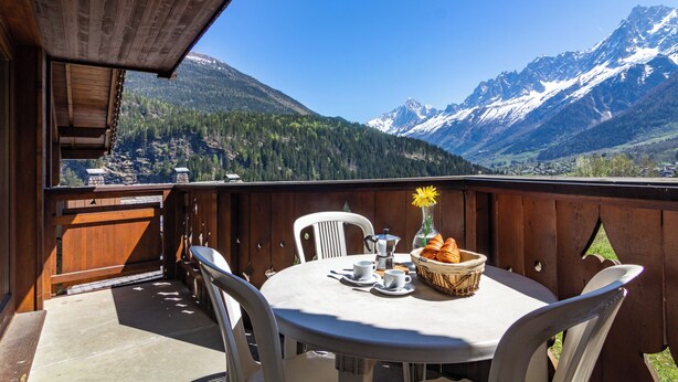 Una terraza de madera con una mesa redonda y sillas, una cesta de croissants, un florero con una flor amarilla y vistas a montañas nevadas.