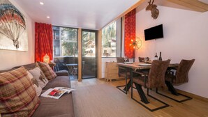 A modern living room with a sofa, dining table, and chairs, a flat-screen TV, and a large window with a view of trees and buildings. - Grand Roc B22 260m from Slopes, View, Parking & WiFi, Argentière (Chamonix), France (Argentière (Chamonix))