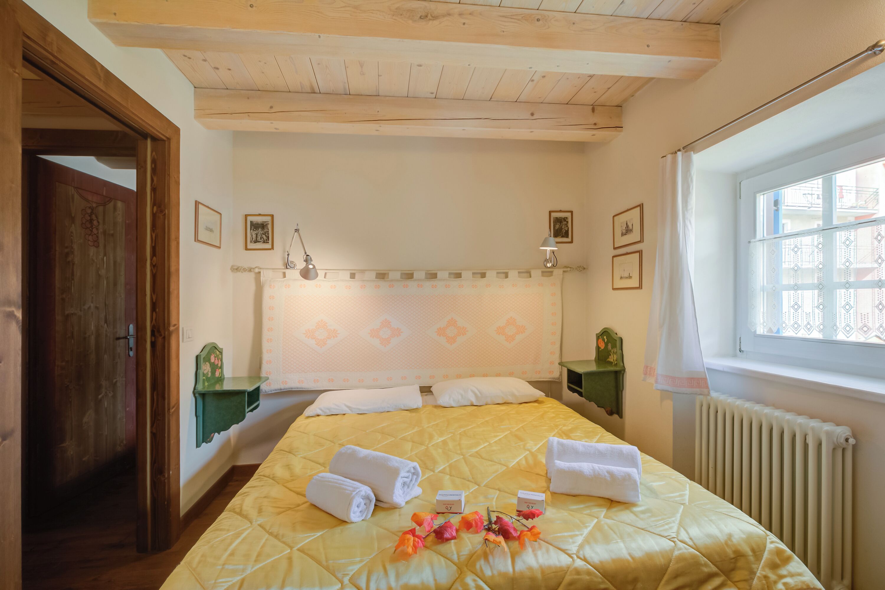 Een slaapkamer met een bed, kussens, een nachtkastje met een lamp, een raam met gordijn en een houten deur.