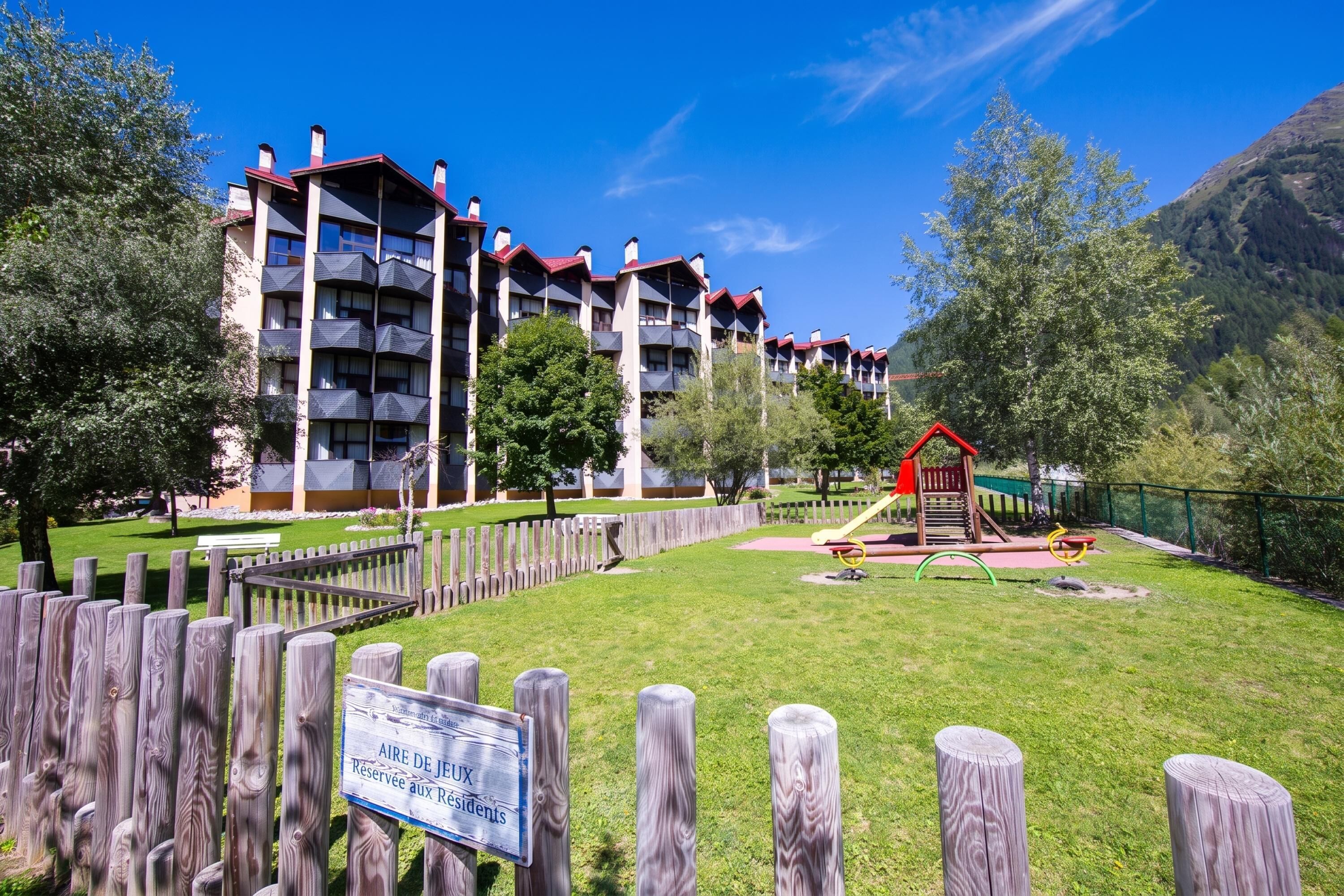 Een meerlaags hotel met balkons, een speeltuin en een omheinde tuin.