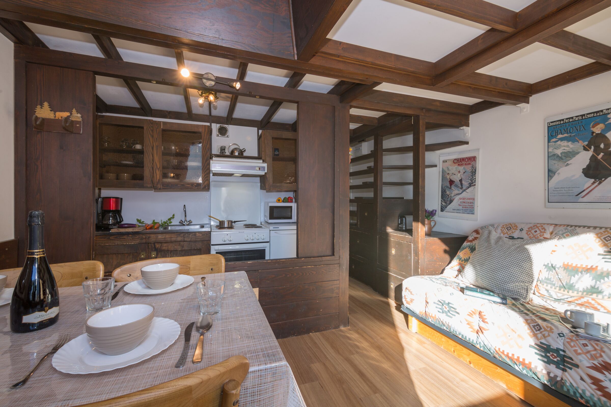 Un coin repas chaleureux avec une table en bois dressée pour deux personnes, un canapé et une cuisine en arrière-plan.