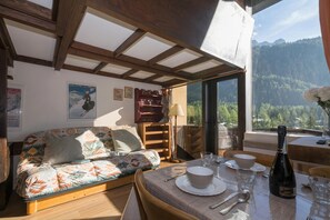 Une pièce chaleureuse avec un plafond en bois, un canapé agrémenté de coussins à motifs, une table à manger avec des assiettes et des verres, et une bouteille de vin.