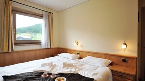 A hotel room with a bed, a bedside table, a nightstand, a window with curtains, and a view of the outdoors.