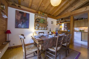 Un coin repas en bois, avec une table dressée pour quatre personnes, entourée de chaises. Une étagère fixée au mur expose différents plats et un tableau.