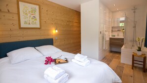 A hotel room with a wooden headboard, a bed with white linens, a wooden side table with a tray of bread and flowers, and a bathroom with a shower and sink.