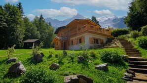 A wooden chalet with a balcony, surrounded by a lush garden and a mountain backdrop.