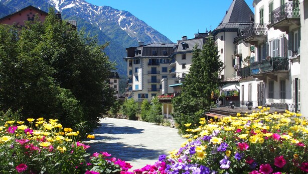 A hotel with a mountain backdrop, a river in the foreground, and colorful flower beds.