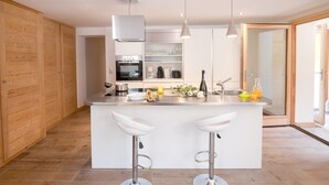 A modern kitchen with a central island, two bar stools, a built-in oven, and a coffee machine.