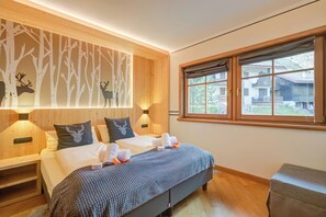 A room with two beds, a large window, wooden walls, and a forest-themed wall decoration.