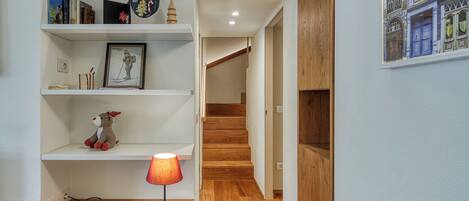 Un pasillo con suelo de madera, una estantería con libros y un cuadro enmarcado, una lámpara sobre una mesa de madera, y una escalera que conduce a otra habitación.
