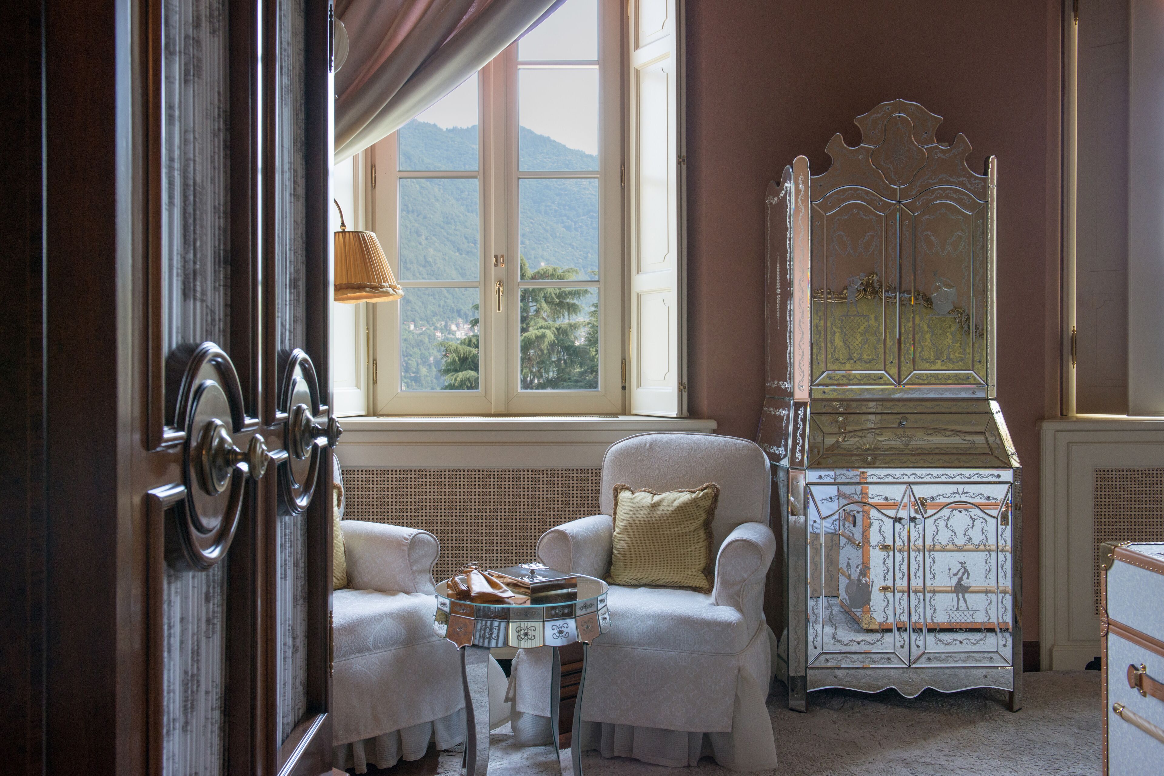 A room with a large window, a white armchair with a cushion, a small table with a tray, and a decorative cabinet.