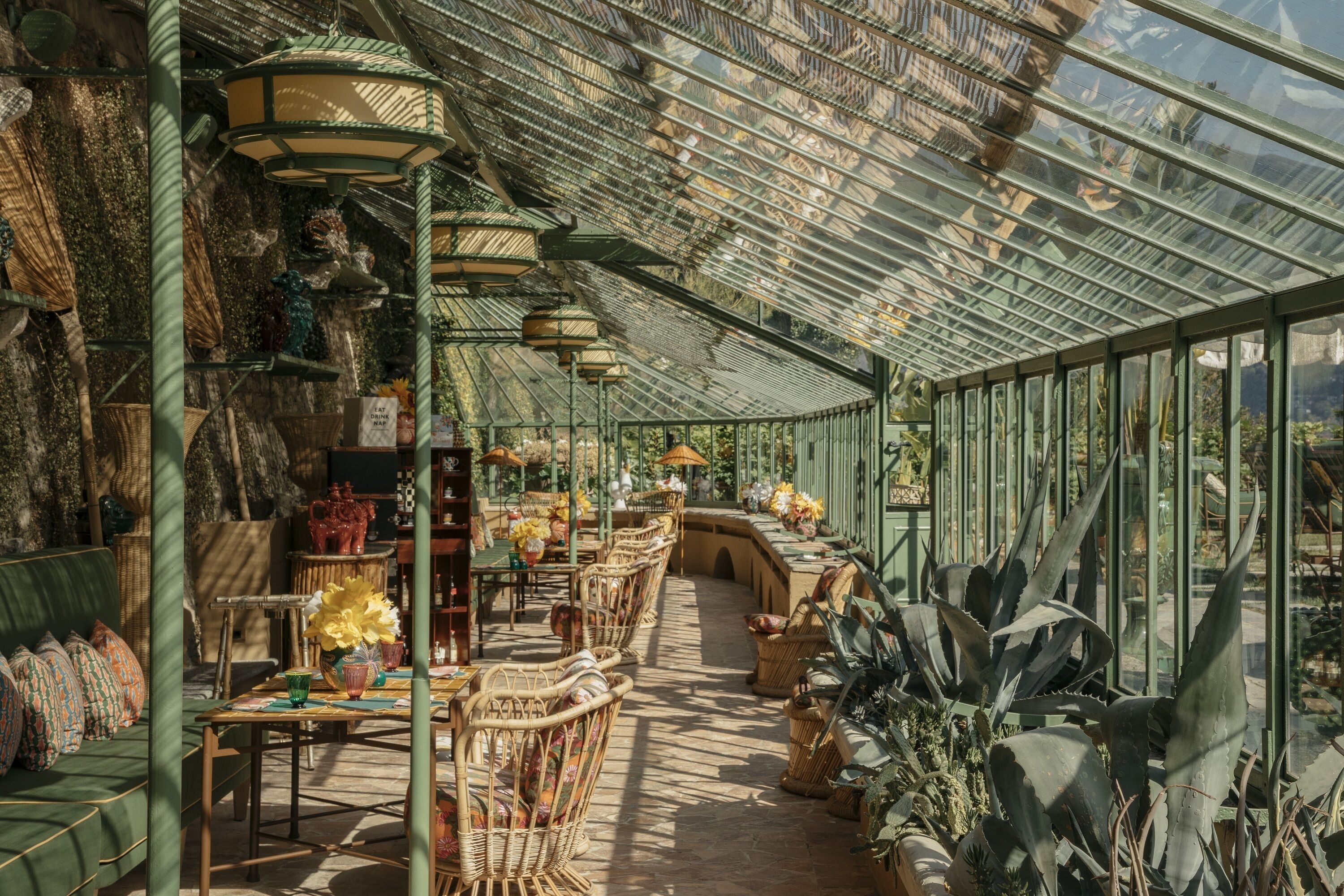 A greenhouse with rows of wicker furniture, potted plants, and hanging lamps.
