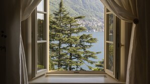 A window with white curtains, a view of a lake, and a forested mountain in the background. - Passalacqua (Moltrasio)