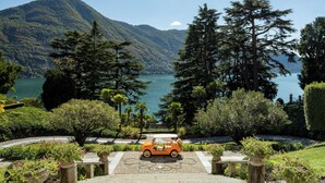 A scenic view of a lake with mountains in the background, a pathway leading to a parked orange car, and lush greenery. - Passalacqua (Moltrasio)