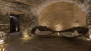 Um quarto com paredes de pedra, duas áreas de estar acolchoadas e um candeeiro.