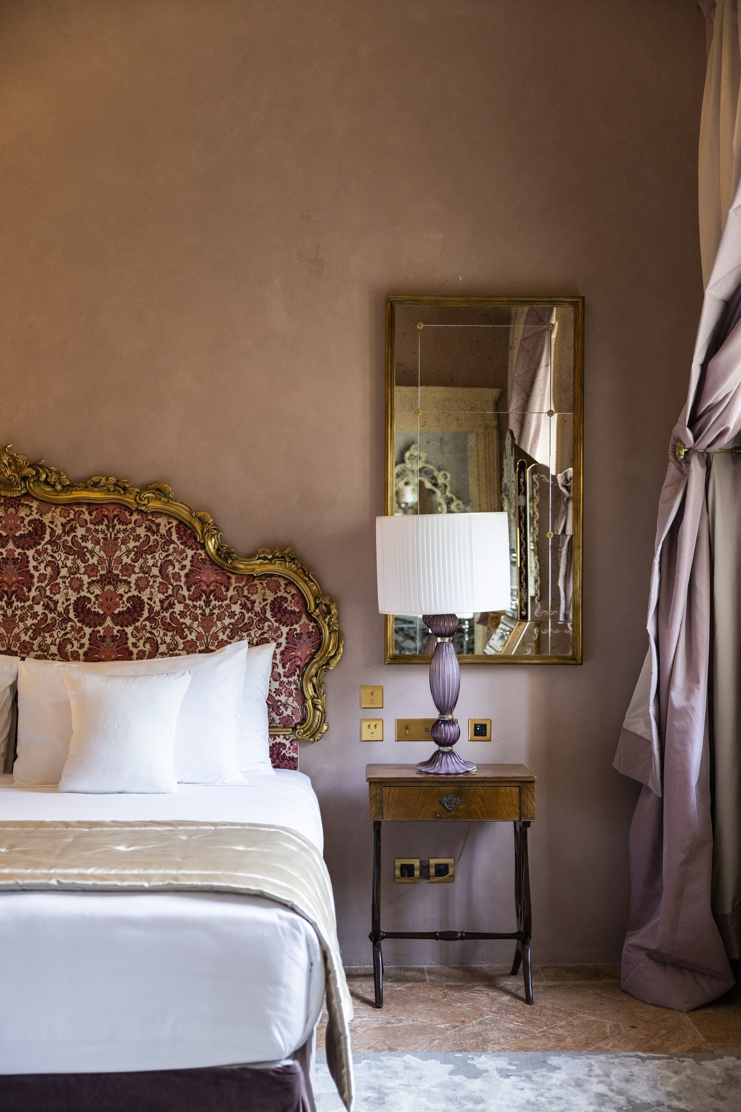 A hotel room with a large bed, a bedside table with a lamp, a mirror, and a curtain.