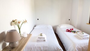 Une chambre d’hôtel avec deux lits simples, une table de chevet avec une lampe, un vase avec des fleurs et une petite étagère avec un livre et une tasse.