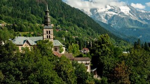 Une église dotée d’un clocher imposant, entourée d’arbres verdoyants et avec une chaîne de montagnes en toile de fond.