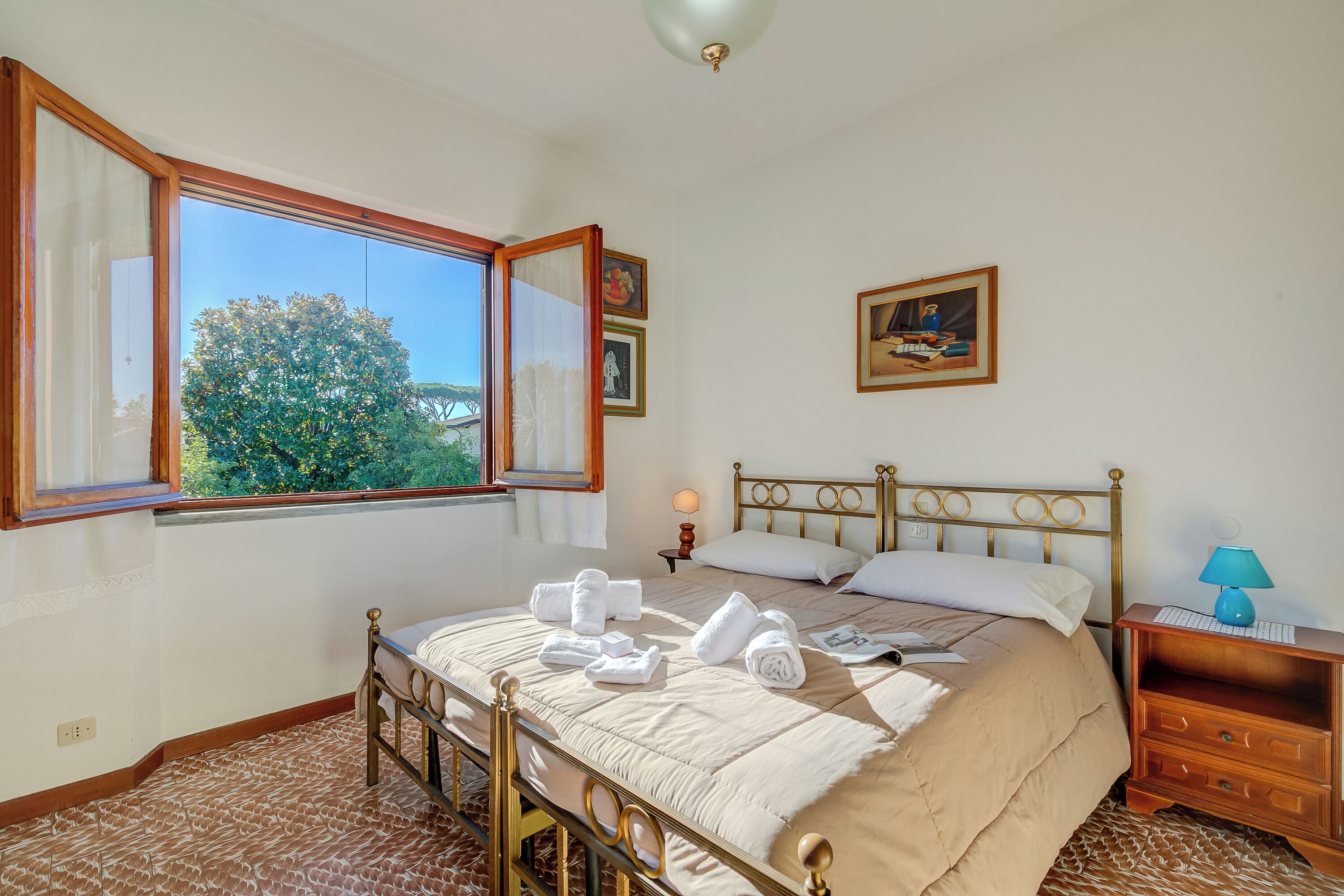 Un dormitorio con un ventanal grande, una cama con toallas, una mesita de noche con una lámpara y un cuadro en la pared.