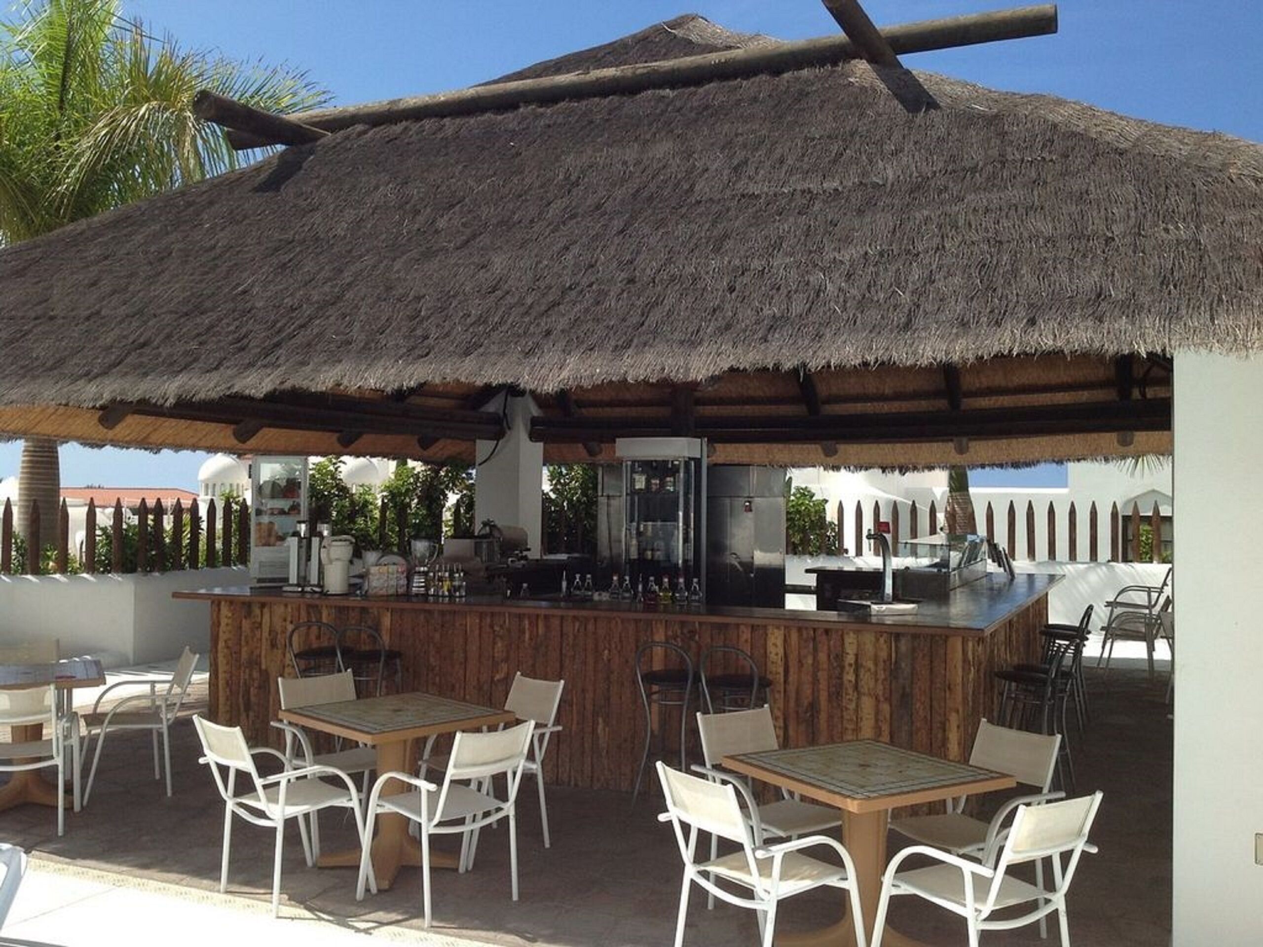 Un espace bar extérieur doté d’un toit de chaume, d’un comptoir en bois et de chaises en métal blanc.
