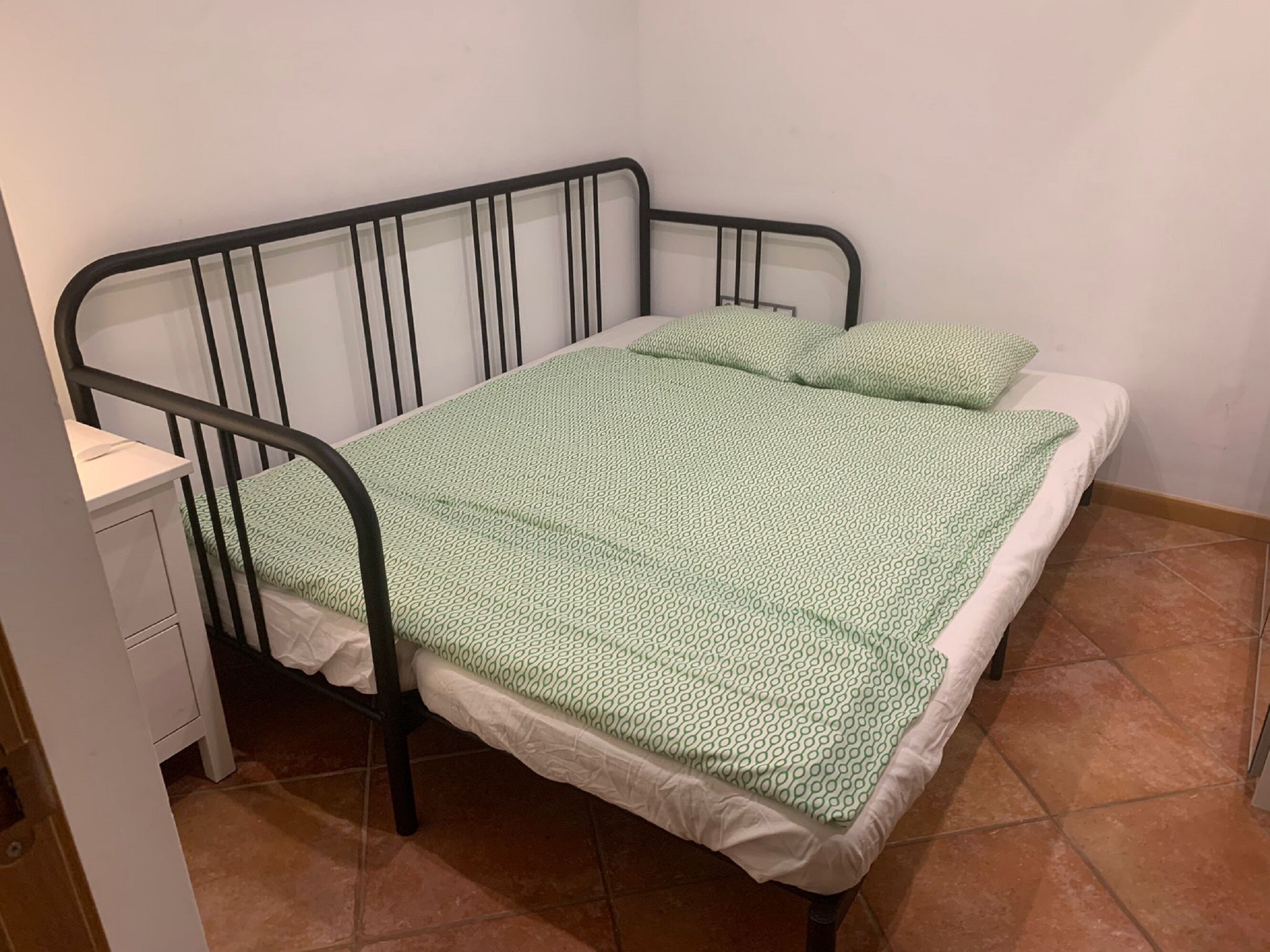 Uma cama de solteiro com estrutura de metal, colchão branco e manta verde estampada.