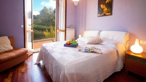Camera da letto con un letto, un comodino, una lampada da lettura, una finestra vista su piante e un quadro appeso al muro.