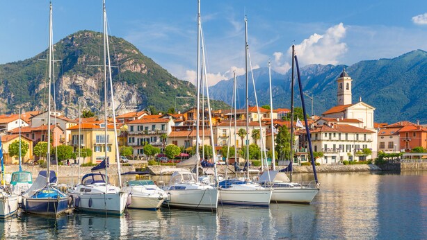 Un porto turistico con diverse barche a vela ormeggiate, una montagna sullo sfondo e una cittadina con edifici e una torre.