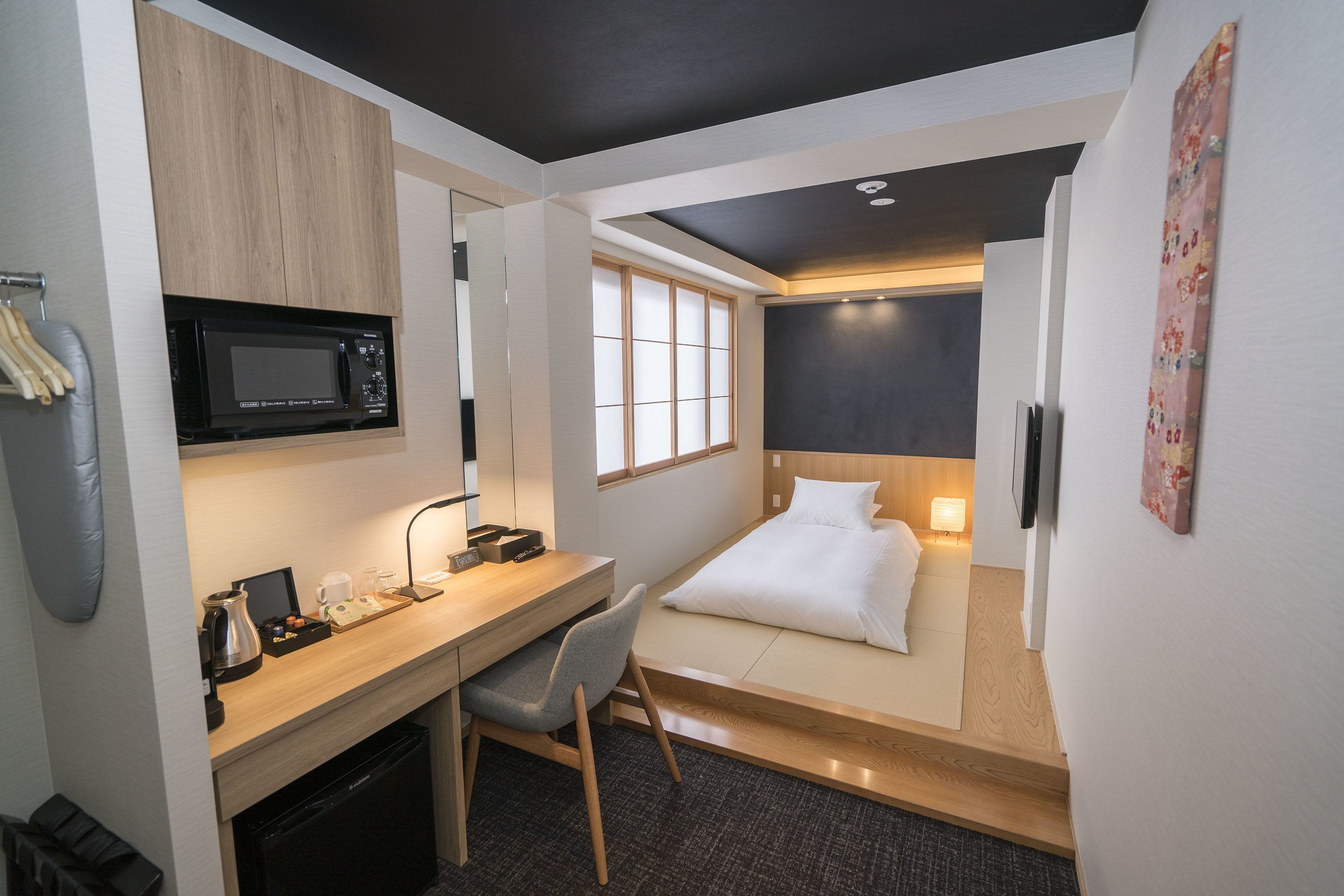 A compact hotel room with a kitchenette, a bed, a desk with a chair, and a window.
