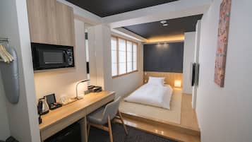 A compact hotel room with a kitchenette, a bed, a desk with a chair, and a window.