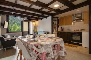 Ein Essbereich mit Tisch für vier Personen, eine Küche mit modernen Geräten und Blick auf einen Wald durch ein großes Fenster.