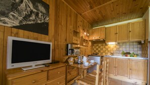 A wooden interior with a flat-screen TV on a wooden dresser, a kitchen area with wooden cabinets, a small refrigerator, and a dining table with chairs.