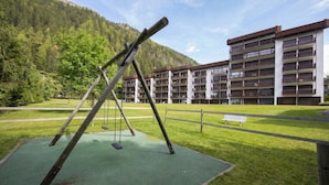 A playground with swings and a bench, surrounded by a grassy area and a multi-story building with balconies.