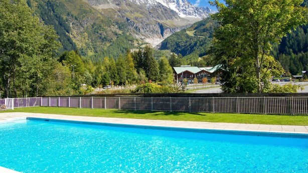 A swimming pool with a mountain backdrop, a fenced area, and a building with a green roof.