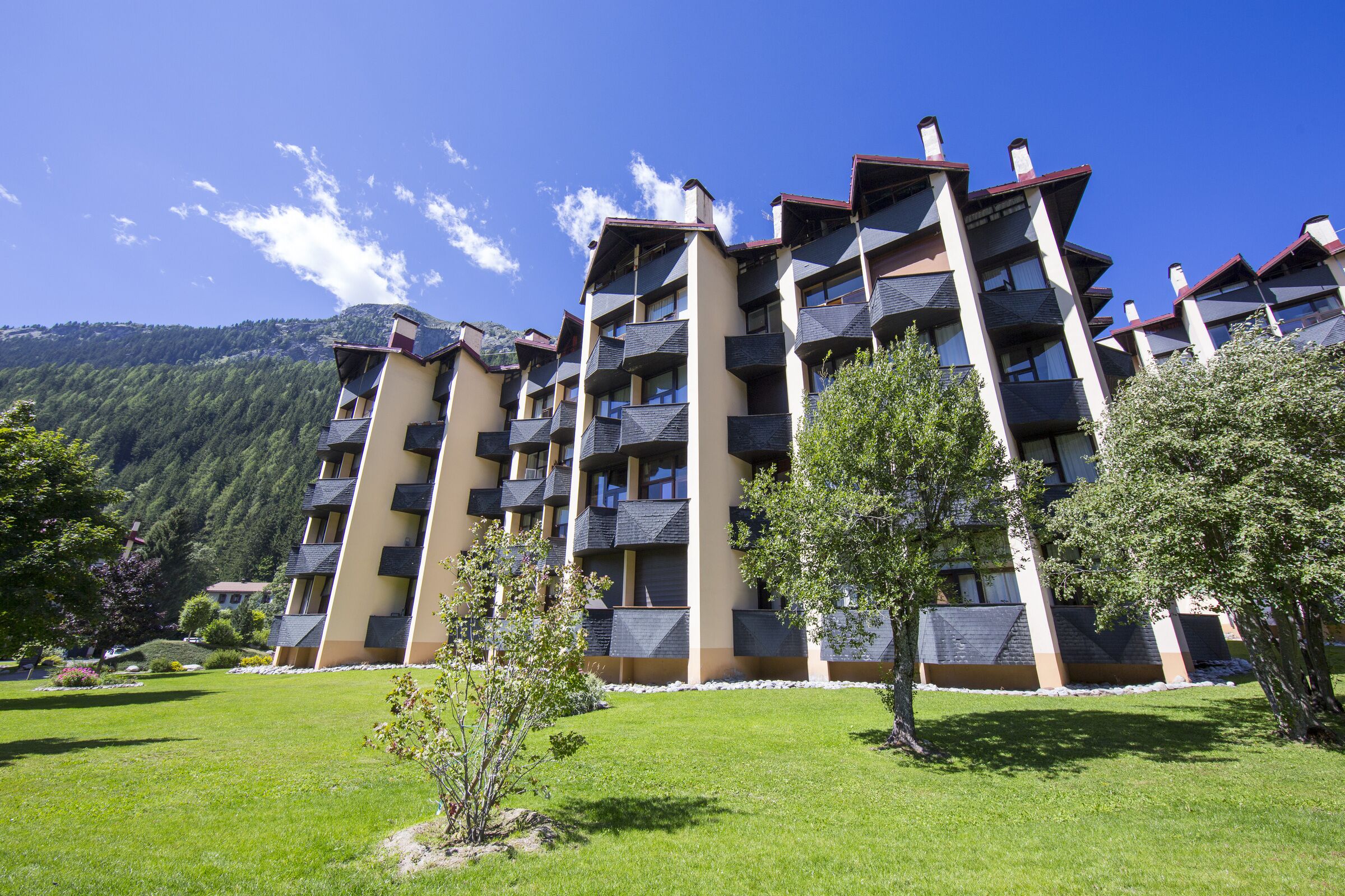 A multi-story hotel with balconies, surrounded by green grass and trees, with mountains in the background.