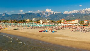 Ein Strand mit Sonnenschirmen und Booten, im Hintergrund Berge und in der Nähe Gebäude.