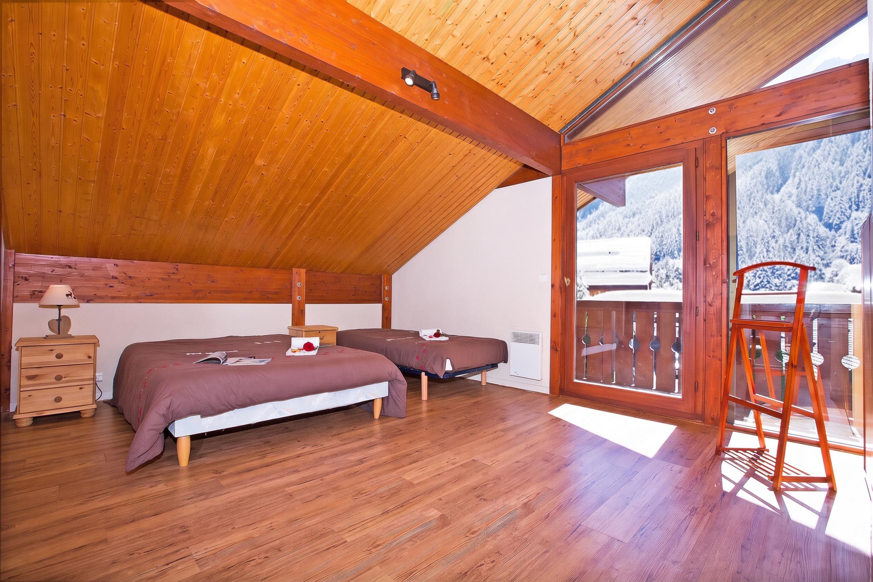 Un dormitorio acogedor con dos camas, suelo de madera, una mesita de noche y un balcón con vistas a montañas nevadas.