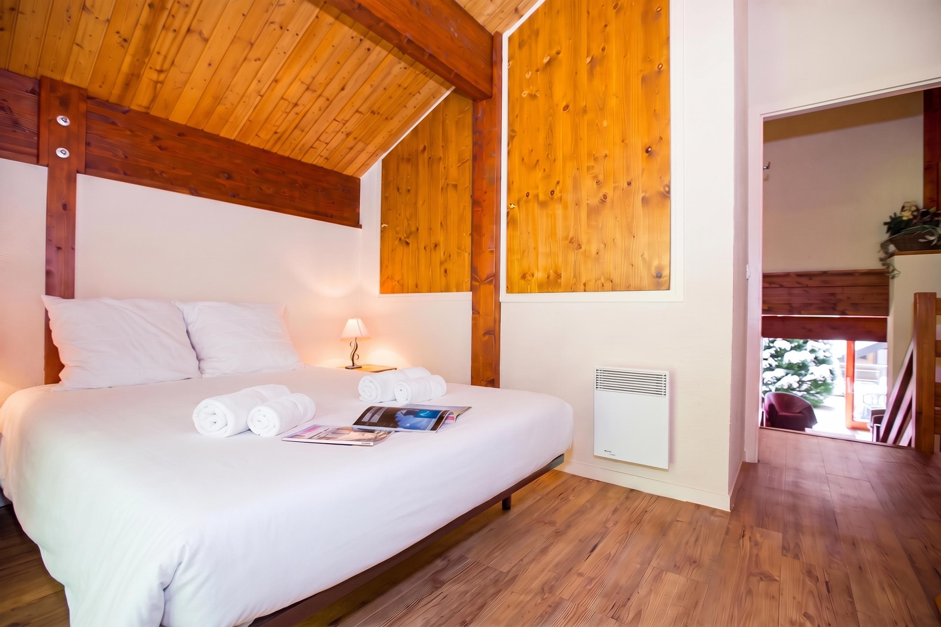 Een slaapkamer met een houten plafond, een bed met witte lakens, een nachtkastje met een lamp en een wandradiator.