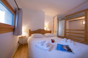Ein ordentlich bezogenes Bett mit aufgerollten Handtüchern und einer Zeitschrift, eine Nachttischlampe und ein Fenster mit Vorhängen.