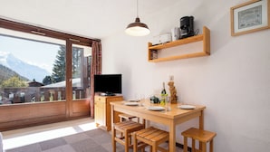 A room with a wooden dining table, chairs, a TV, a shelf with items, and a window with a mountain view. - La Verte 12 Ski In/Ski Out | Grands Montets | Argentière Chamonix, Argentière (Chamonix), France (Argentière (Chamonix))