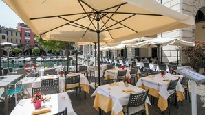 Un área de comedor al aire libre con mesas y sillas, una piscina al fondo, y un edificio con un cartel que dice "HOTEL".