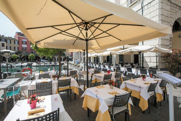 An outdoor dining area with tables and chairs, a pool in the background, and a building with a sign that reads "HOTEL". - Residenza Verde - 3 Pools & Garage, Desenzano Del Garda, Italy (Desenzano Del Garda)