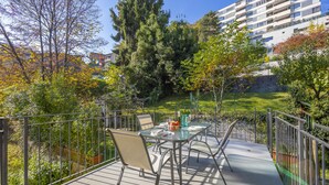 Un balcón con una mesa y sillas de vidrio, rodeado por una barandilla de metal, con vistas a un jardín con árboles y un edificio al fondo.