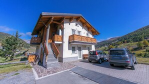 A two-story building with a wooden balcony, a staircase, and a parking area with cars. - Cervo Ski in - Ski out 200m, Livigno, Italy (Livigno)