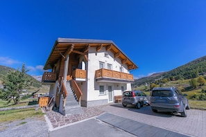 A two-story building with a wooden balcony, a staircase, and a parking area with cars.