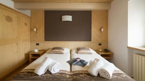 A double bed with a mountain-patterned bedspread, two bedside tables with lamps, a wooden wardrobe, and a plain wall above the bed. - Rubino Ski in, Livigno, Italy (Livigno)