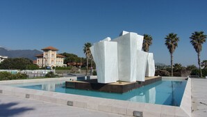 Un espace piscine moderne, agrémenté d’une sculpture blanche originale qui en constitue l’élément central, et entouré de palmiers sous un ciel d’un bleu limpide.