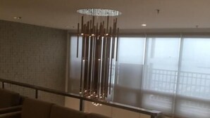 Un salon moderne avec un canapé, un luminaire suspendu et une vue sur l’extérieur grâce à de grandes fenêtres.