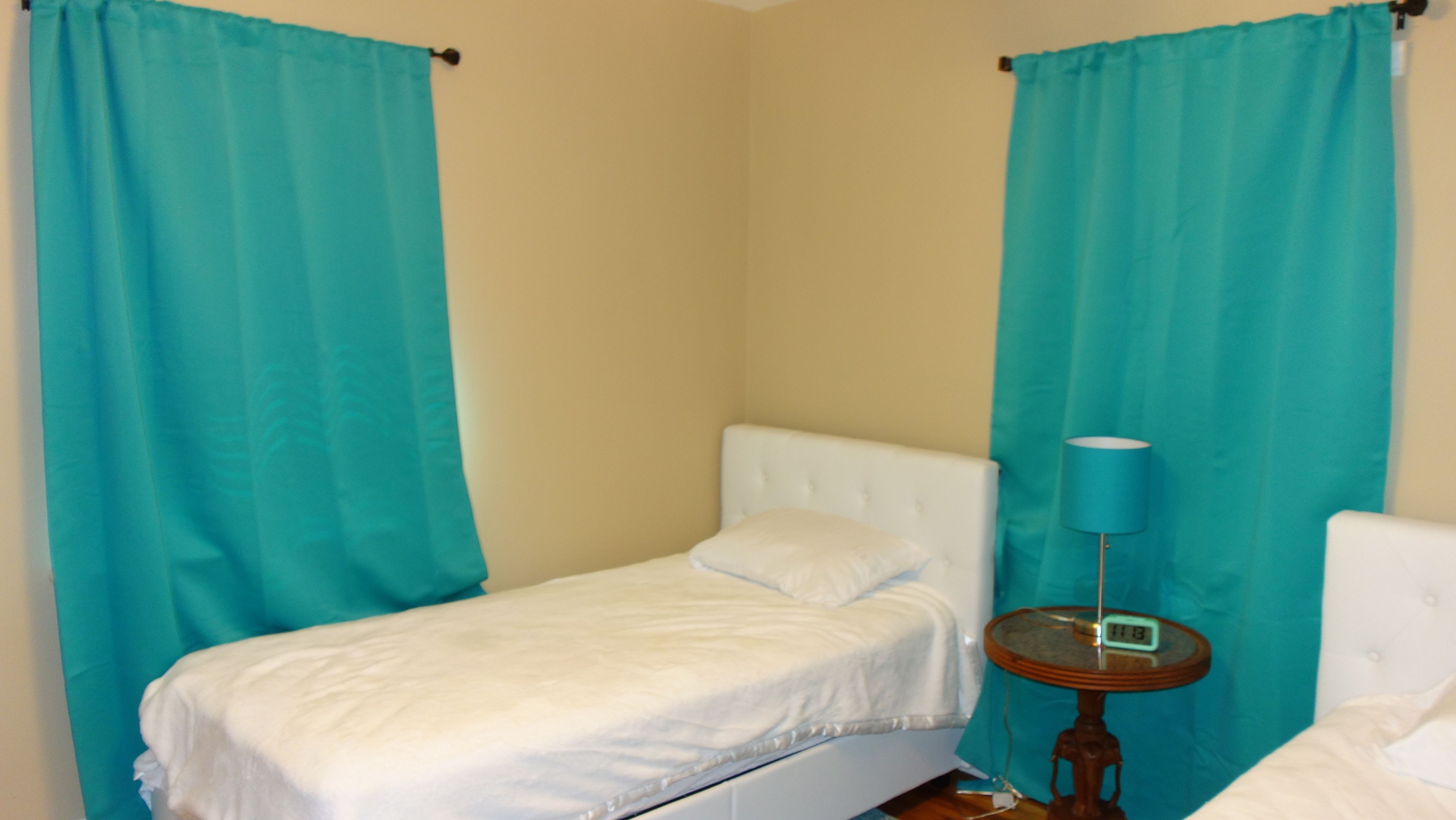 A small, single-bed room with teal curtains, a bedside lamp, and a small table with a digital clock.